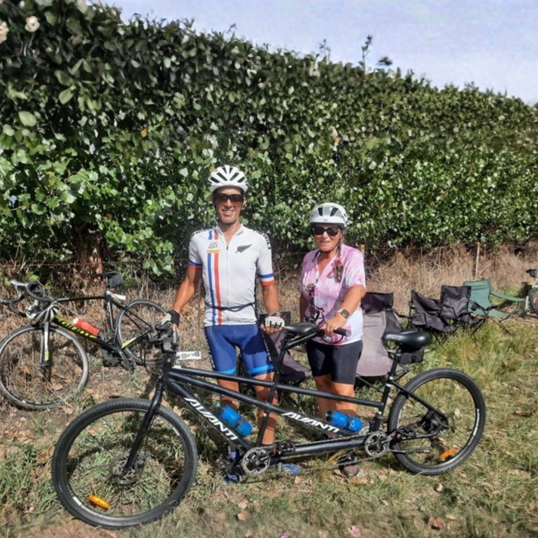 A man and woman in cycling attire with a tandem bicycle in an outdoor setting.