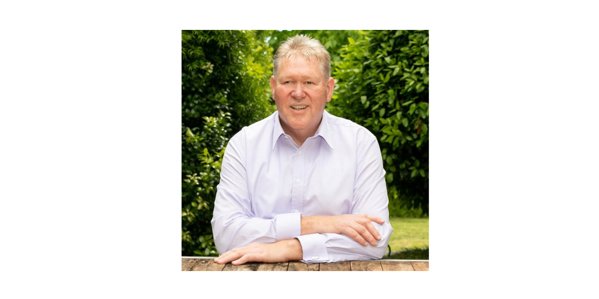 Professional portrait of a smiling man with strawberry-blonde hair wearing a light lavender button-down shirt, sitting with arms crossed at a wooden table against a lush green hedge background.
