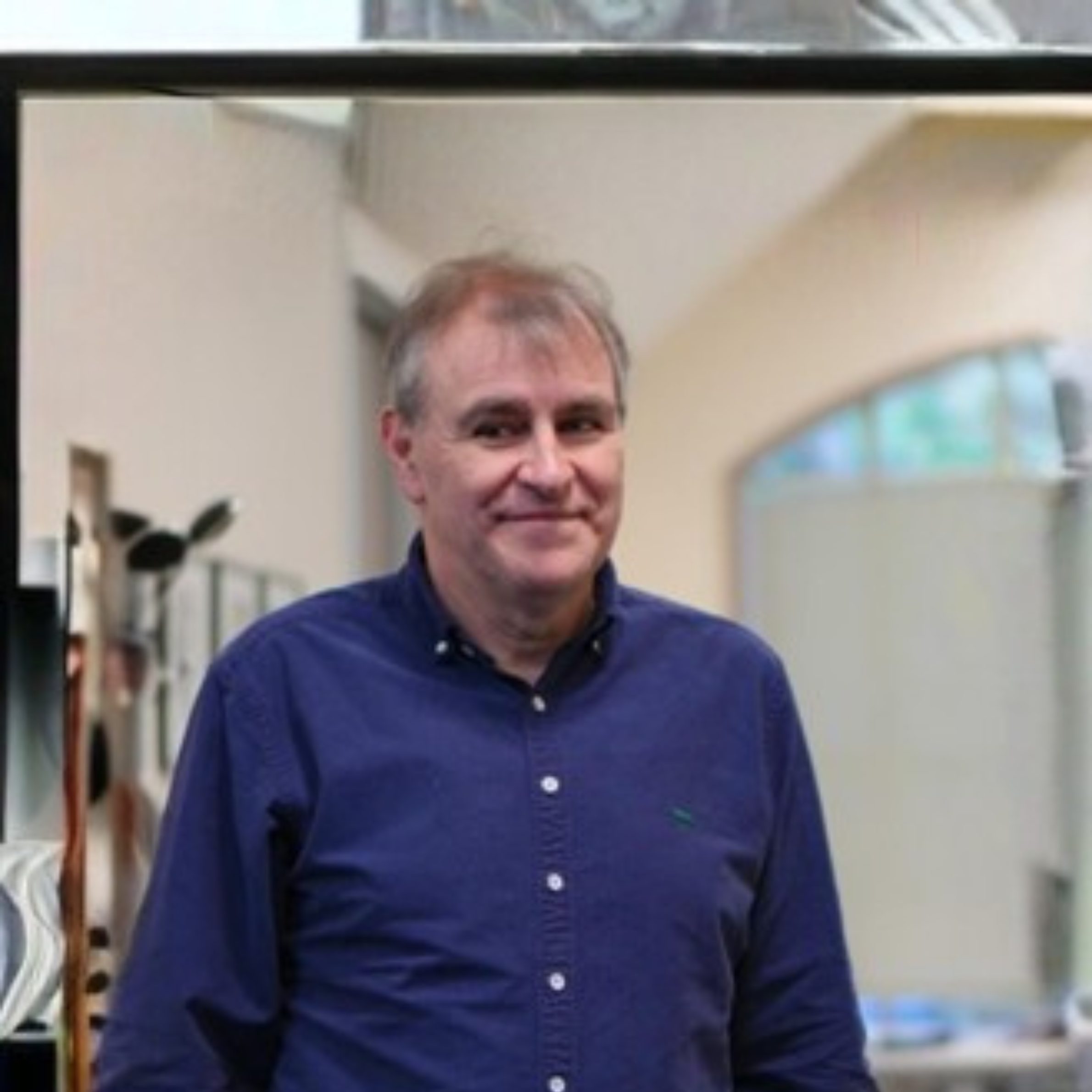 A middle-aged man with grey hair and a dark blue shirt standing in an office environment.