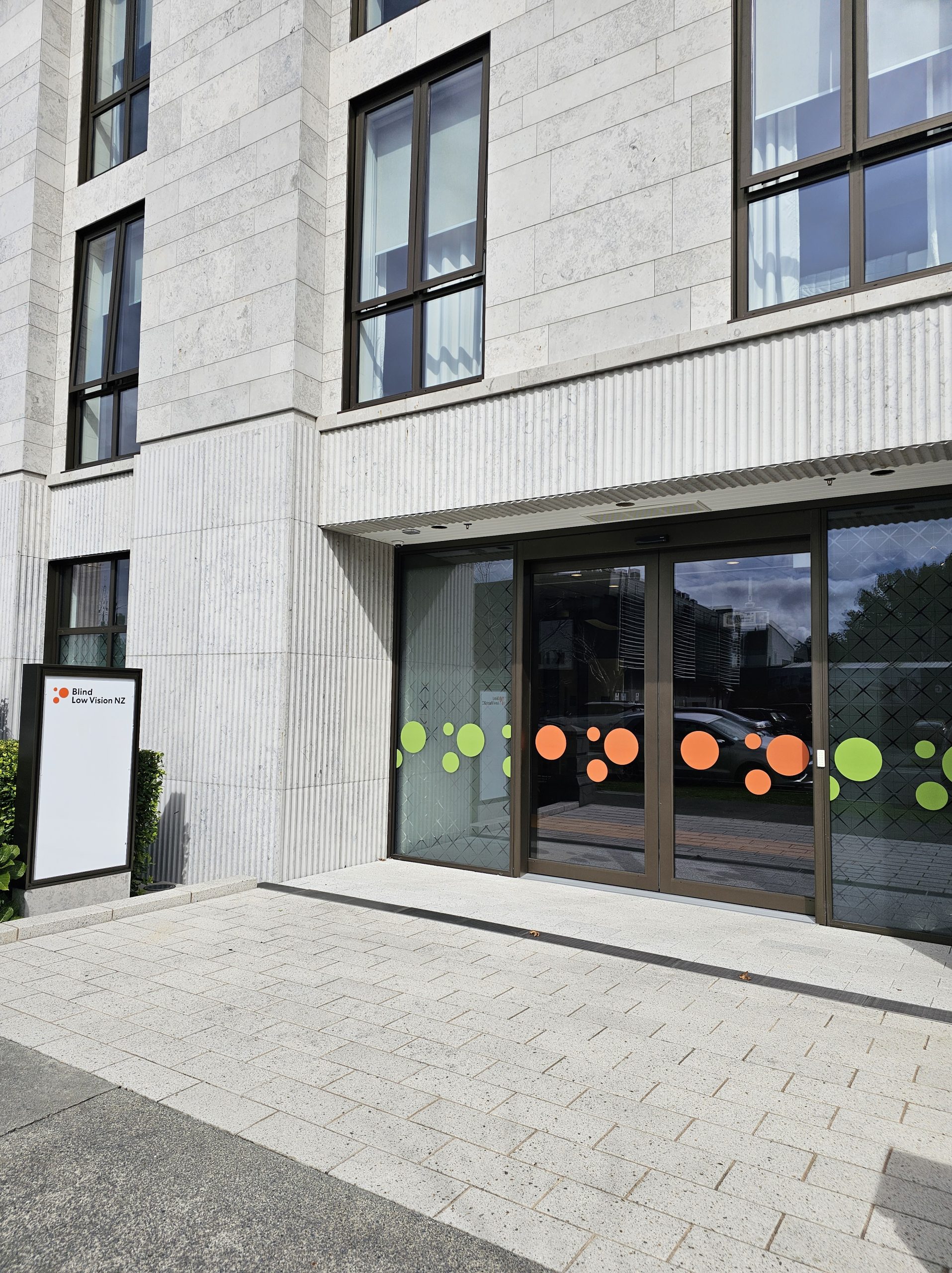The outside of a building with the sign Blind Low Vision NZ. The entrance feature automatic sliding doors with the Blind Low Vision Logo on the front in both orange and green.