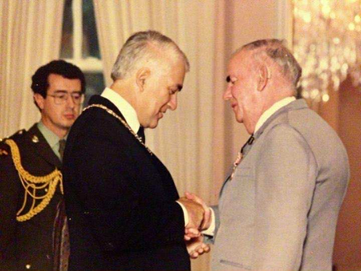 Two men in a formal setting, shaking hands ceremoniously. On the left is Sir Paul Reeves and on the right is Terry Small.